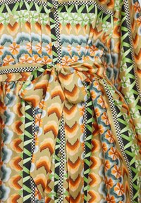 Gros plan sur un tissu aux motifs géométriques et floraux vibrants en orange, vert, noir et beige, présentant un nœud noué et un devant boutonné.
