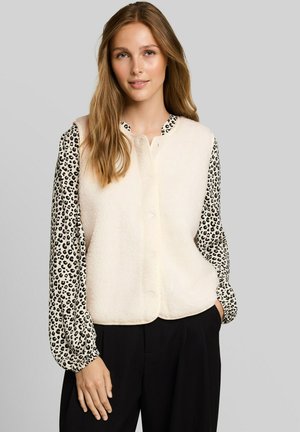 Jeune femme aux longs cheveux châtain clair portant une veste en polaire blanche par-dessus un chemisier à manches longues imprimé léopard et un pantalon noir.