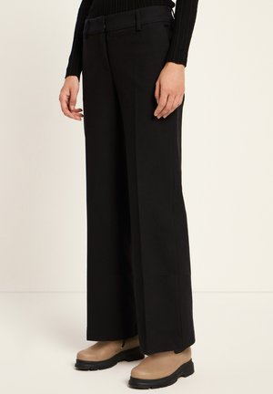 Person in schwarzen weiten Hosen, einem schwarzen gerippten Pullover und beigen ankle boots mit schwarzen Sohle vor einem hellen Hintergrund.