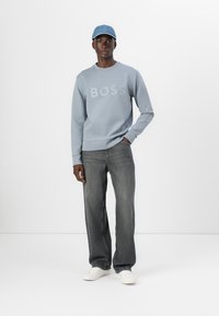 Un giovane uomo si trova di fronte a uno sfondo semplice, indossando una felpa blu chiaro con la scritta "BOSS", jeans grigi e sneakers bianche.