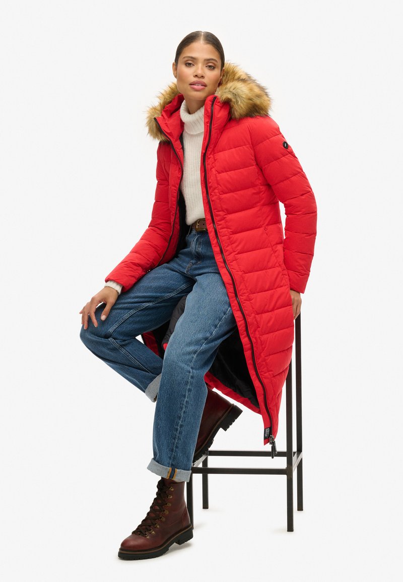 Rode gewatteerde jas met een met bont gevoerde capuchon, ritssluiting en zijzakken, gedragen over een wit sweater en blauwe jeans. Bruine veterschoenen eronder.