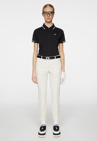 Black short-sleeve polo shirt with white accents, paired with cream patterned pants, black sneakers, and clear sporty glasses. White glove on left hand.