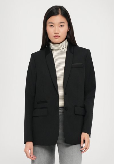 Black tailored blazer with satin-lapel, two front pockets, and single button closure. Worn over a striped turtleneck, paired with gray pants.