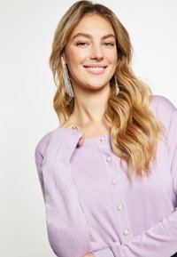 Lila kardigán, puha anyagból készült, gombos záródással, díszes kerek díszítményekkel és bordázott mandzsettákkal. Hosszú, hullámos haj.