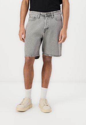Mann trägt graue Jeansshorts, weiße Socken und beige Sneaker und steht vor weißem Hintergrund.