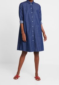 Jeansklänning i mörkblått, med knappfront, långa ärmar som är uppvikta vid ärmsluten, en enda bröstficka och en lös silhuett.