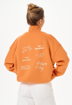 Femme de dos portant un sweat-shirt orange avec des phrases positives comme « crée ta vie », « sois sans peur » et « rêve grand » imprimées dans le dos.