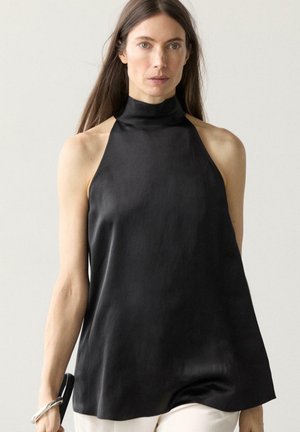 Frau mit langen dunklen Haaren, die ein ärmelloses schwarzes Satinoberteil mit hohem Kragen trägt und ein silbernes Armband am linken Handgelenk hält.