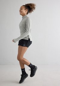 Haut à manches longues gris clair avec une fermeture éclair et des fentes sur les côtés, associé à un short noir et des baskets noires. Les chaussettes présentent un logo swoosh blanc.
