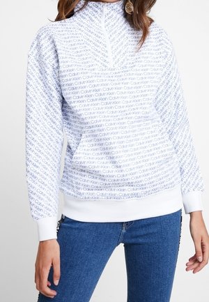 Sudadera blanca con patrón de logotipos de Calvin Klein en azul, cuello alto, cierre de cremallera y puños de canalé. Combinada con jeans azules.