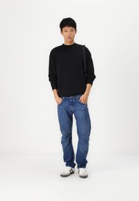 Black sweatshirt, blue cargo jeans with multiple pockets, and white sneakers with black accents. Model stands in a neutral background.
