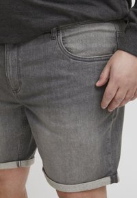 Sivi traper šortsevi s preklopljenim rubom, s blagim izblijeđenjem i klasičnim dizajnom s pet džepova. Prikaz izbliza s rukom oslonjenom na tkaninu.
