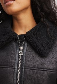 Veste en cuir noir avec une finition texturée, fermeture éclair à l'avant et un col en shearling noir pelucheux, mettant en valeur des matériaux et des designs contrastés.