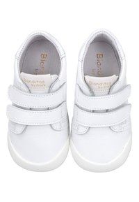 Zapatillas sintéticas blancas con dos correas de Velcro, parte superior texturizada, punta redondeada y suela de goma. Marca visible en el interior del zapato.