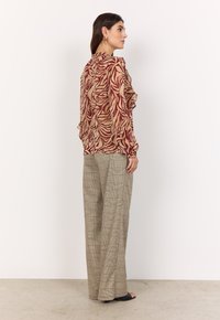 Blusa fluida con un motivo astratto rosso e beige, abbinata a pantaloni a gamba larga a quadri in crema, grigio e dettagli rossi.