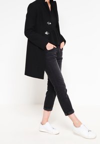 Kvinna i svart rock och jeans, med händerna i fickorna, står med ett ben böjt, vita sneakers på fötterna, mot en vit bakgrund.