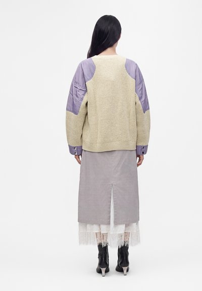 Femme aux longs cheveux foncés porte un pull en maille beige avec des patchs violets, une jupe midi grise, un jupon en dentelle blanche et des bottes à talons noires, vue de dos.