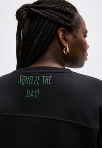 Frau mit geflochtenen Haaren, die goldene Creolen und ein schwarzes Oberteil mit grünem Schriftzug "SQUEEZE THE DAY!" am oberen Rücken trägt.