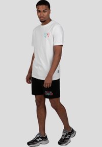 Camiseta de algodón blanca con mangas cortas que presenta letras coloridas en el lado izquierdo del pecho. Combinada con pantalones cortos negros y zapatillas deportivas.