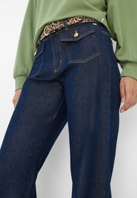 Dunkelblaue Denim-Jeans mit einer lockeren Passform, ausgestattet mit zwei Gesäßtaschen und goldenen Hardware-Details. Kombiniert mit einem grünen Sweatshirt und einem Gürtel im Leopardenmuster.