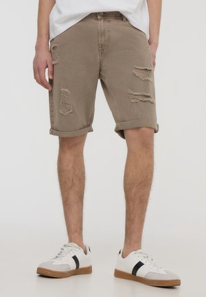 Uomo che indossa pantaloncini beige strappati con orli arrotolati e sneakers bianche con strisce nere, in piedi davanti a uno sfondo semplice.
