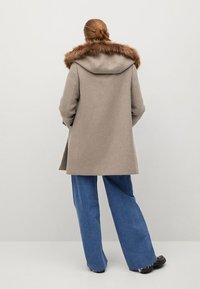 Abrigo de lana beige con capucha de piel, que presenta un corte holgado y una abertura en la parte posterior, combinado con pantalones de mezclilla azul de pierna ancha.