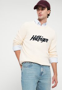 Κρεμ κεντημένο πουλόβερ με λογότυπο "Hilfiger", συνδυασμένο με ανοιχτό μπλε τζιν παντελόνι. Το μοντέλο φορά επίσης ένα μπορντώ καπέλο και ένα μπλε ριγέ πουκάμισο.
