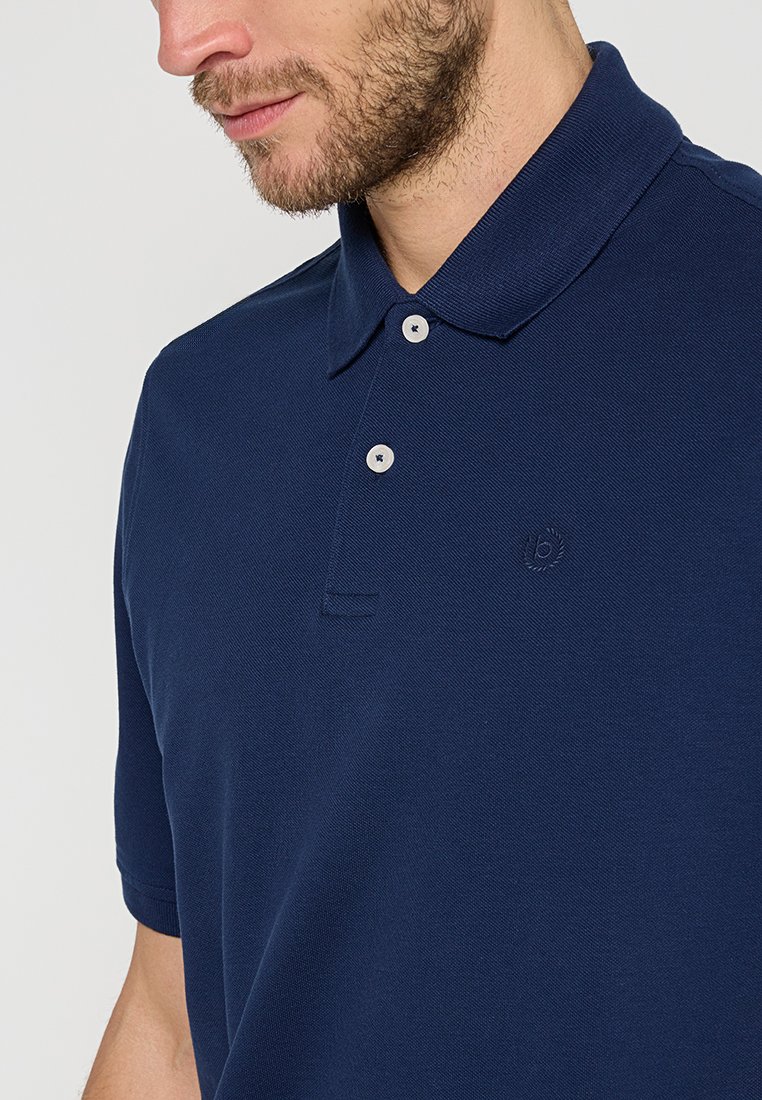 Homme portant un polo bleu marine à manches courtes avec deux boutons blancs et un petit logo brodé sur la poitrine.