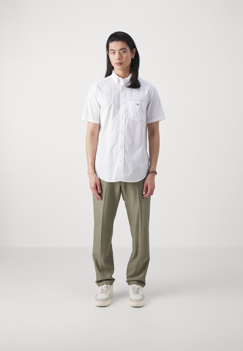 Chemise blanche à manches courtes avec fermeture à boutons et poche, assortie à un pantalon vert olive. Le modèle porte des baskets crème. Design simple et épuré.