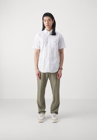 Young person with long black hair stands wearing a white short-sleeve shirt, light olive pants, and white sneakers against a plain background.