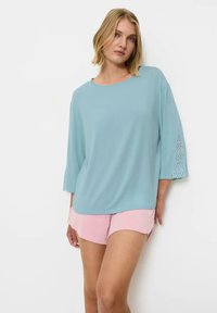 Jasnoniebieska luźna bluzka z rękawami do trzech czwartych, ozdobiona koronkowymi detalami. Noszona z pastelowymi różowymi szortami. Gładka tekstura, casualowy fason.