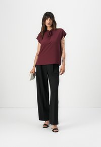 Burgundy kortermet bluse med knytehals; kombinert med svarte vide bukser og hælsandaler, holdt en metallisk clutch.