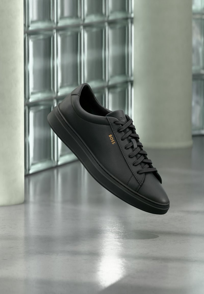 Baskets basses en cuir noir avec lacets noirs et logo BOSS doré sur le côté, flottant au-dessus d’un sol gris brillant dans un espace intérieur moderne.