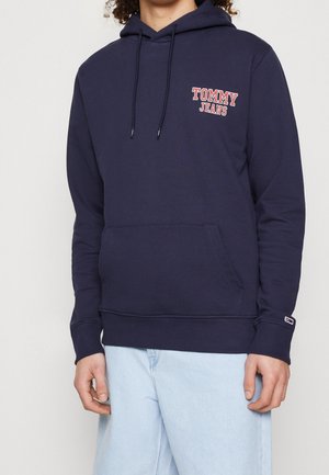 Osoba nosząca granatową bluzę z kapturem z logo "Tommy Jeans" w kolorze czerwonym i białym na piersi oraz jasnoniebieskie jeansy.