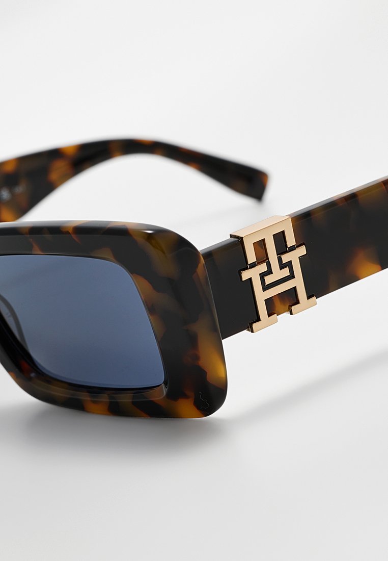 Lunettes de soleil en écaille de tortue avec un design épais, des verres sombres et un accent de logo doré sur la branche. Texture en plastique lisse.