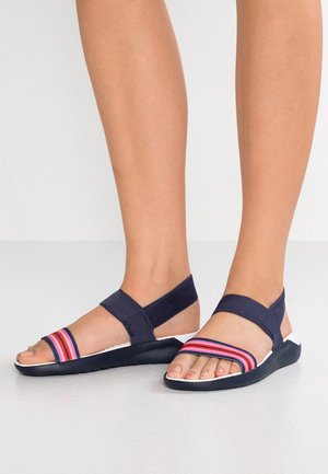 Pieds portant des sandales bleu marine avec des sangles élastiques, avec des sangles avant rayées roses et rouges sur un sol clair.