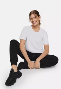 Witte katoenen t-shirt met korte mouwen en een grafische print. Zwarte joggers en zwarte sportieve schoenen met een gestructureerde zool, zittend met de benen gekruist.
