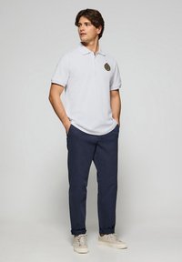 Witte polo met korte mouwen en geborduurd logo, gecombineerd met marineblauwe broek en beige sneakers. De outfit heeft een casual ontwerp.