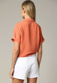 Korte mouwen, textuur oranje blouse met een knopdetail aan de achterkant, gecombineerd met witte shorts met een subtiel patroon.