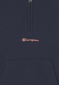 Felpa con zip di colore blu scuro, con logo "Champion" rosa e una tasca frontale, caratterizzata da un tessuto liscio e strutturato.