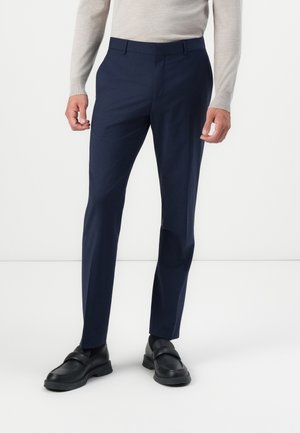Navyblauwe op maat gemaakte broek met een slim fit, gemaakt van een gladde stof. Draagt een lichte trui en zwarte instappers.