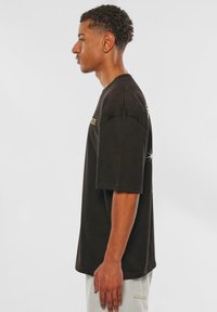 Sort bomulds t-shirt med korte ærmer og en afslappet pasform, med gyldne bogstaver på brystet og et grafisk design på ryggen.