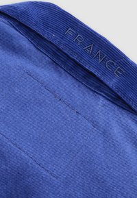 Μπλε ύφασμα με ραβδωτό γιακά κεντημένο με τη λέξη "FRANCE" και ραμμένη τετράγωνη επιφάνεια κάτω από τον γιακά σε ανοιχτό υπόβαθρο.