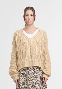 Young woman wearing a beige chunky knit sweater over a white shirt and a leopard print skirt, standing against a plain background.