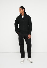 Maglione nero a maglia con colletto alto e dettagli con zip, abbinato a pantaloni cargo neri e scarpe da ginnastica bianche. Texture lisce, vestibilità casual.