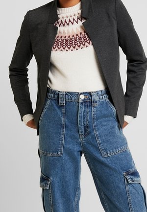 Blazer gris foncé avec col cranté, pull blanc orné d'un motif géométrique rouge et marron, et pantalon cargo en denim bleu taille haute.