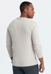 Camicia a maniche lunghe color beige chiaro con scollatura a girocollo, texture morbida, maniche aderenti e un'etichetta logo discreta all'orlo, indossata su uno sfondo neutro.