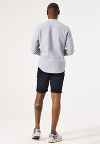 Camisa de manga larga azul claro, pantalones cortos de mezclilla oscuros y zapatillas multicolores. Presenta un tatuaje visible en la pierna inferior.