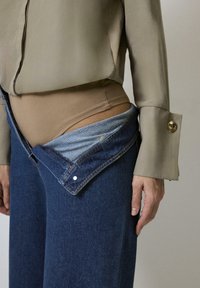 Bodysuit beige dipadukan dengan celana jeans denim biru gelap yang tidak terbutton, dengan ikat pinggang biru muda yang kontras dan atasan lengan panjang berwarna hijau zaitun.