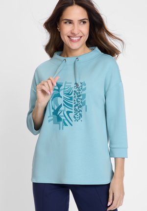 Lächelnde Frau mit langen braunen Haaren trägt einen hellblauen Pullover mit abstrakten tealfarbenen Mustern und marineblauen Hosen und hält einen Hoodie-Zug.